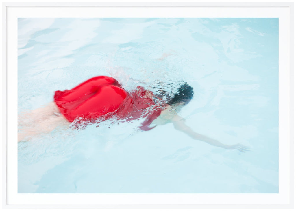 Poster av kvinna i röd klänning dykandes ner i ljusblå pool. Vit ram.