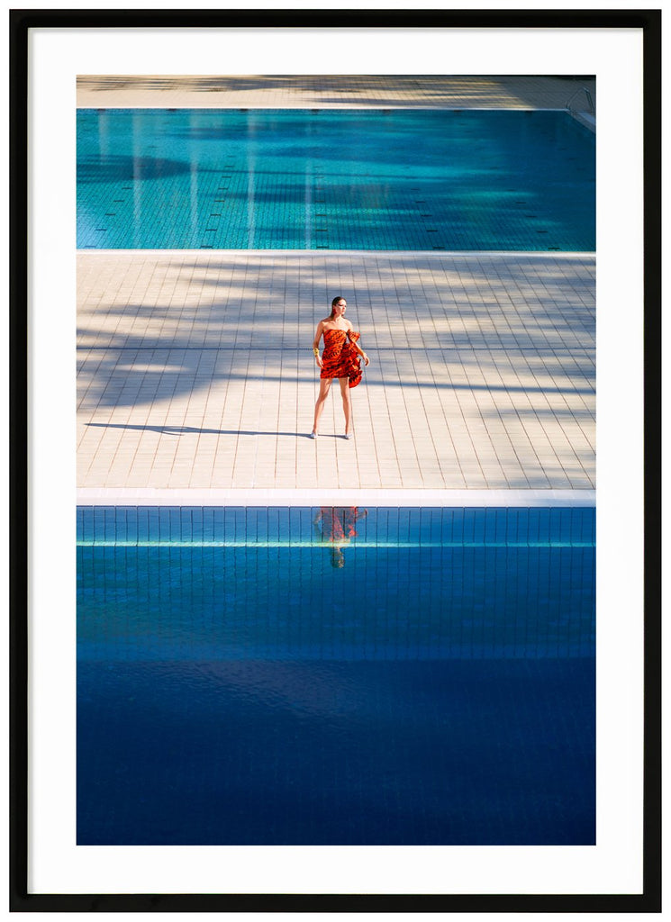 Poster av kvinna i orange klänning som står ensam mellan två stora simbassänger. Svart ram.