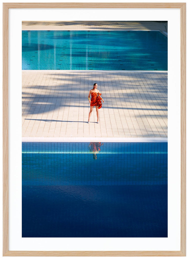 Poster av kvinna i orange klänning som står ensam mellan två stora simbassänger. Ekram.