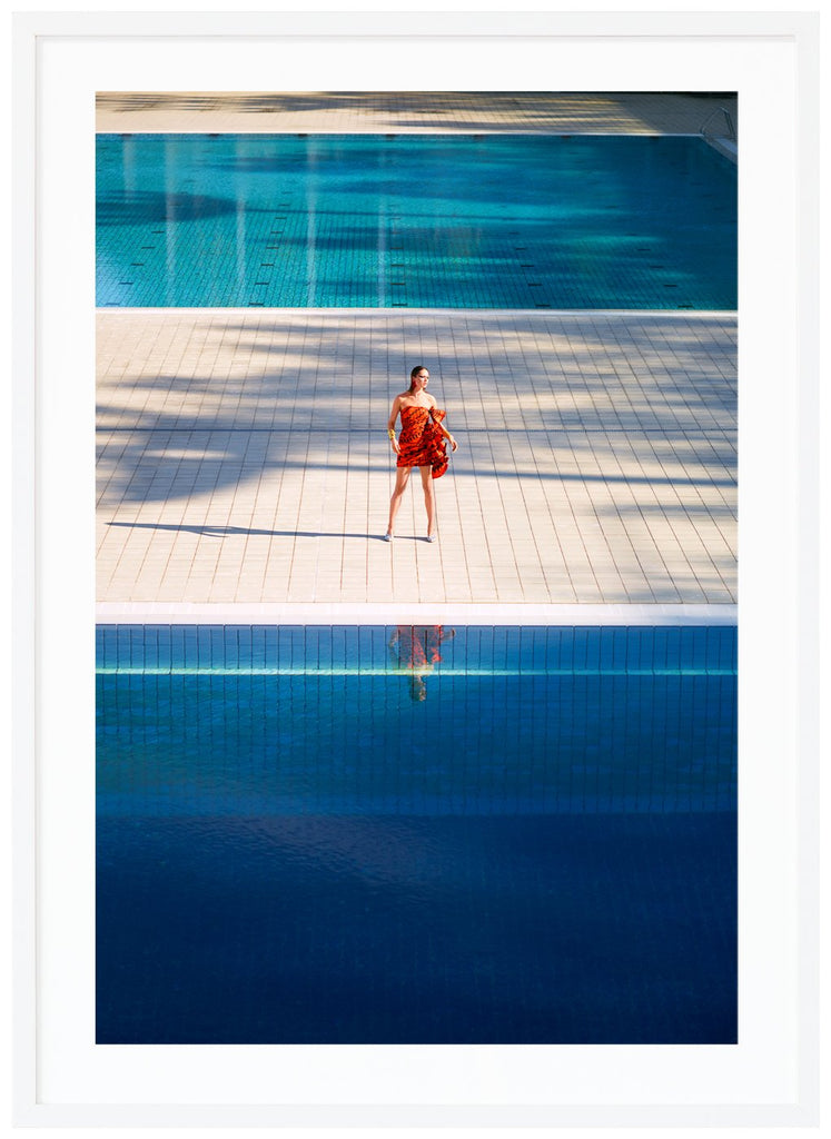 Poster av kvinna i orange klänning som står ensam mellan två stora simbassänger. Vit ram.