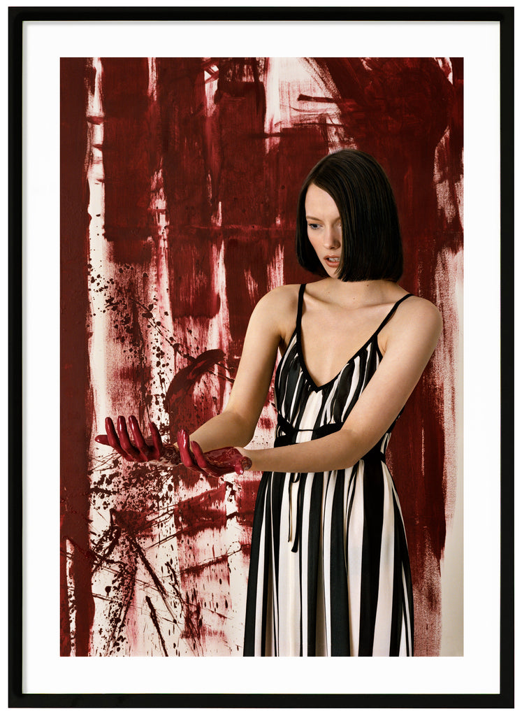 Poster av kvinna ståendes framför konstverk tittandes på sina händer. Svart ram.