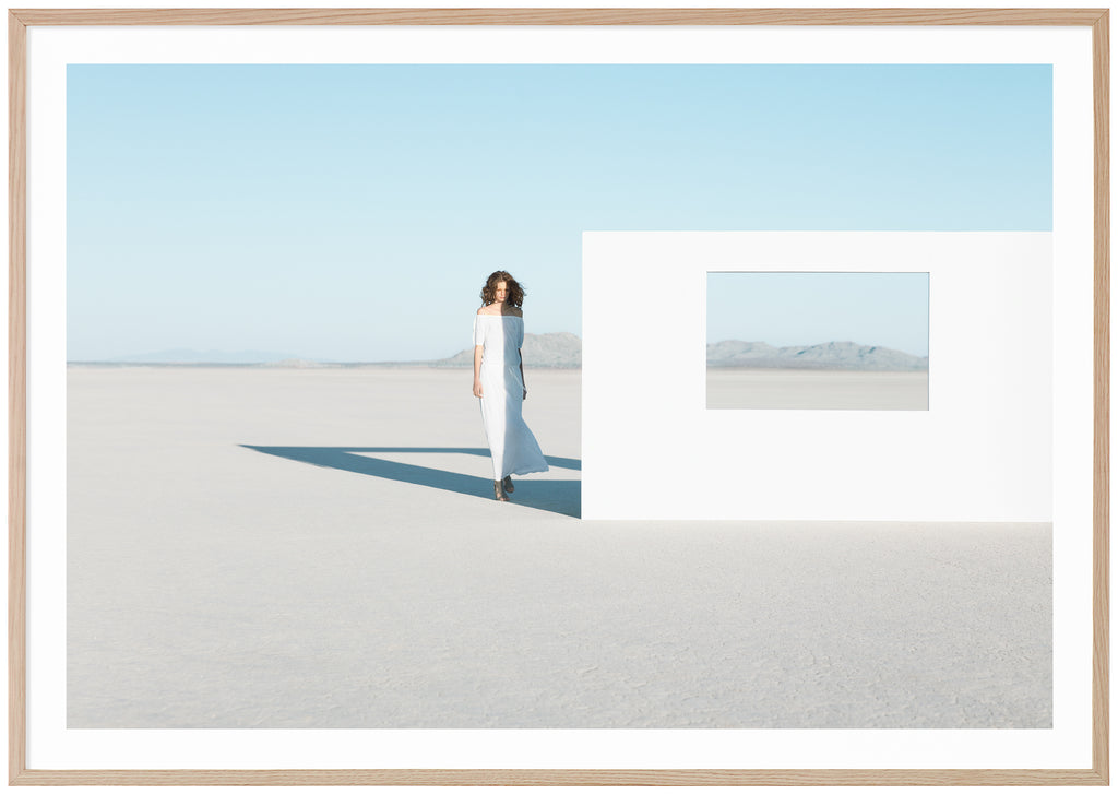 Poster av modefotografi av kvinna i vit klänning i öknen. Står bredvid vit vägg med öppet fönster. Liggande format. Ekram.