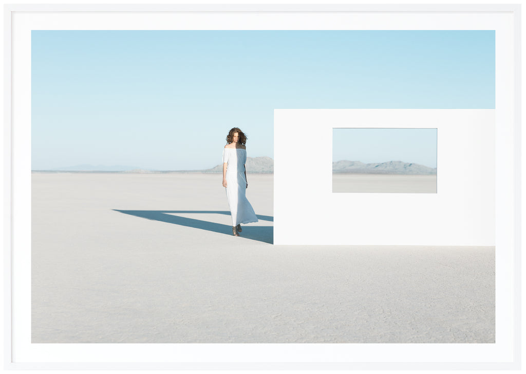 Poster av modefotografi av kvinna i vit klänning i öknen. Står bredvid vit vägg med öppet fönster. Liggande format. Vit ram.
