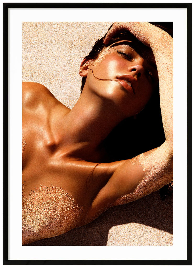 Poster av kvinna liggandes i solen på rygg på sand med handen över pannan. En slinga hår i munnen. Svart ram.
