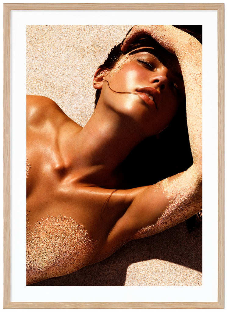 Poster av kvinna liggandes i solen på rygg på sand med handen över pannan. En slinga hår i munnen. Ekram.