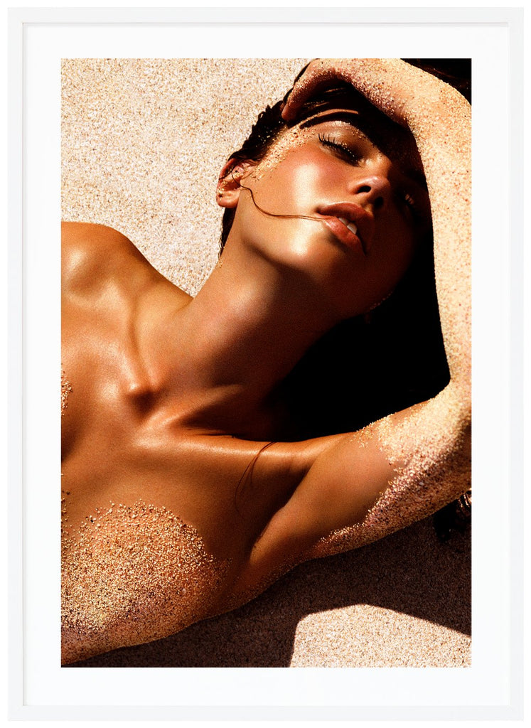 Poster av kvinna liggandes i solen på rygg på sand med handen över pannan. En slinga hår i munnen. Vit ram.
