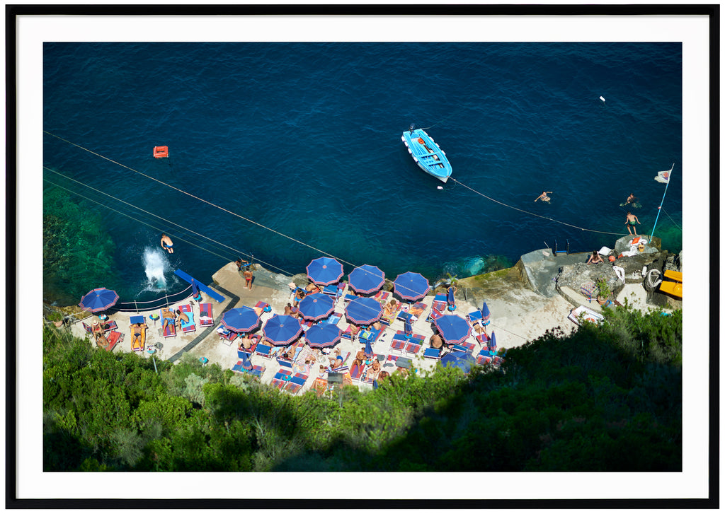 Poster av badplats med blå parasol och vatten. Badande folk och en ljusblå båt. Liggade format. Svart ram.