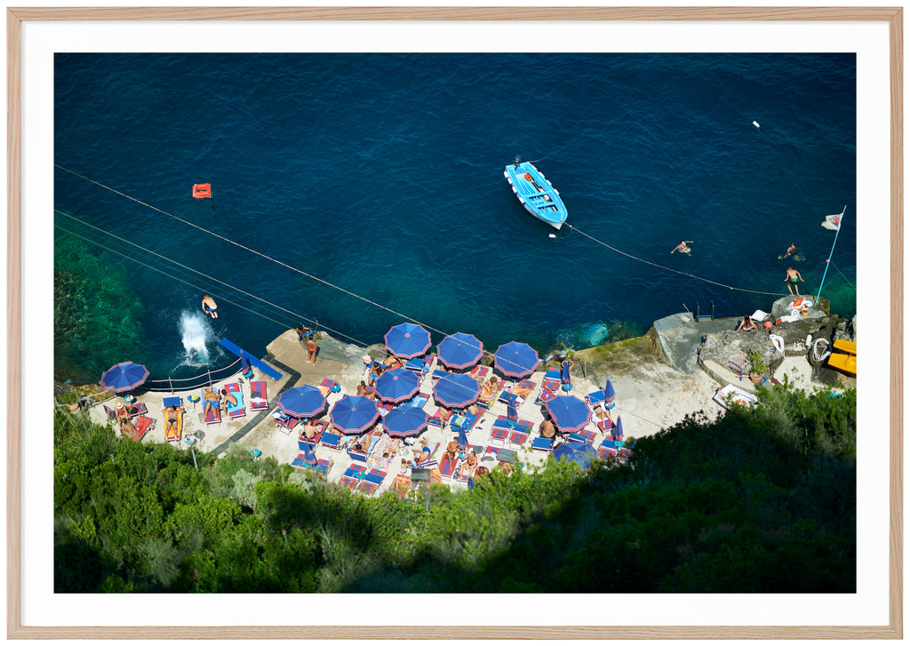 Poster av badplats med blå parasol och vatten. Badande folk och en ljusblå båt. Liggade format. Ekram.