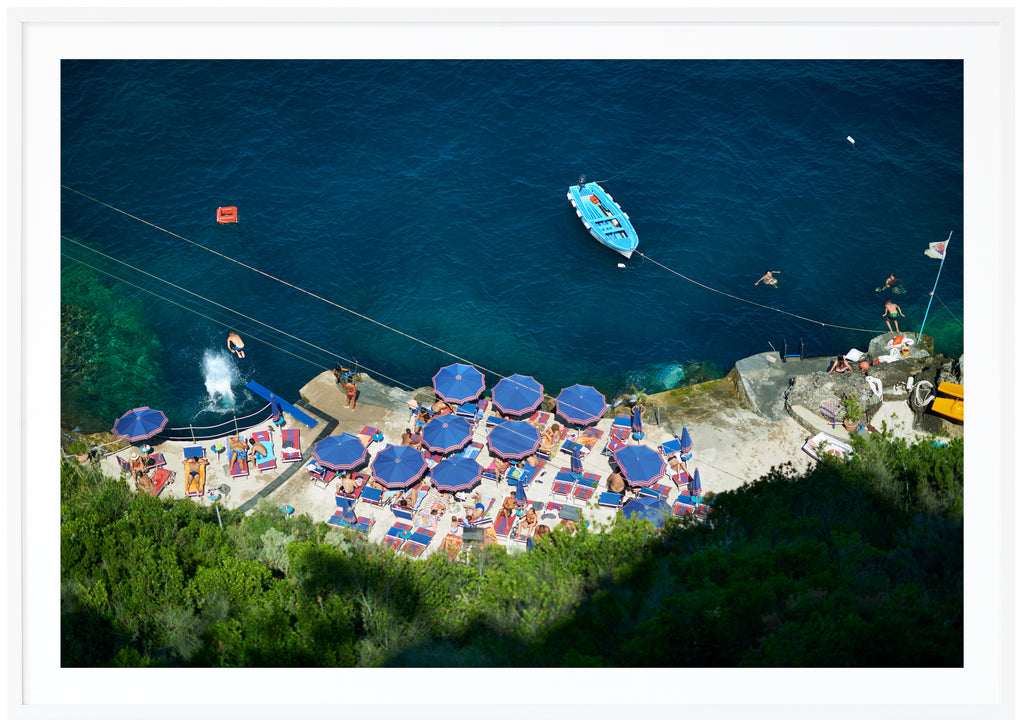 Poster av badplats med blå parasol och vatten. Badande folk och en ljusblå båt. Liggade format. Vit ram.