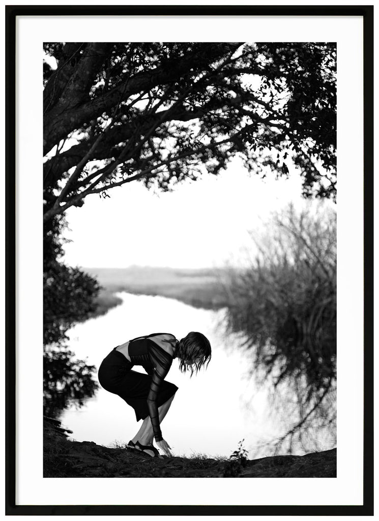 Poster av en kvinna som böjer sig vid vatten. Svart ram.