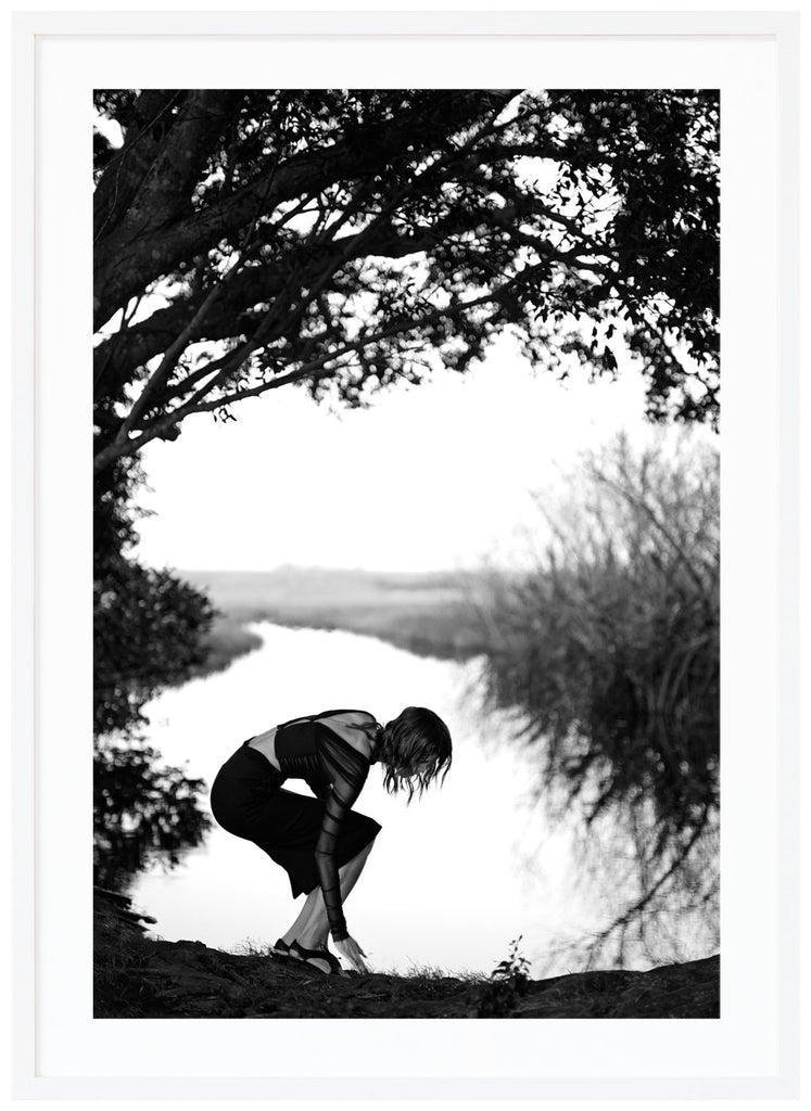 Poster av en kvinna som böjer sig vid vatten. Vit ram.