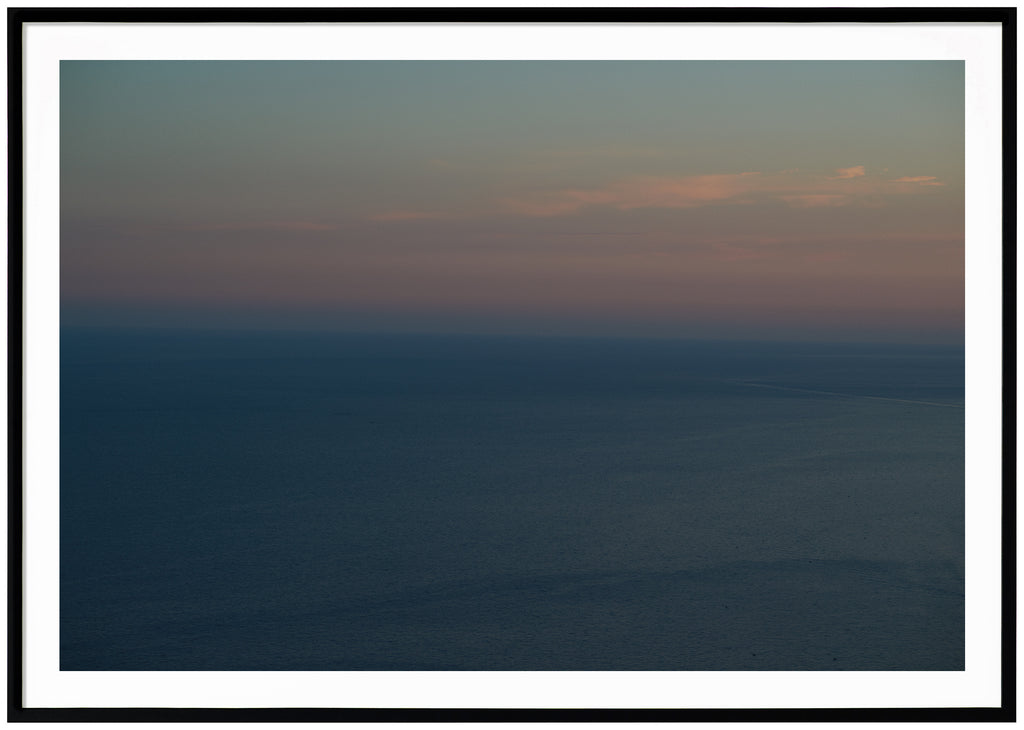 Poster av medelhavet vid Amalfikusten. Himlen är ljusblå orange och rosa i ljusa toner. Havet är blått. Liggande format. Svart ram
