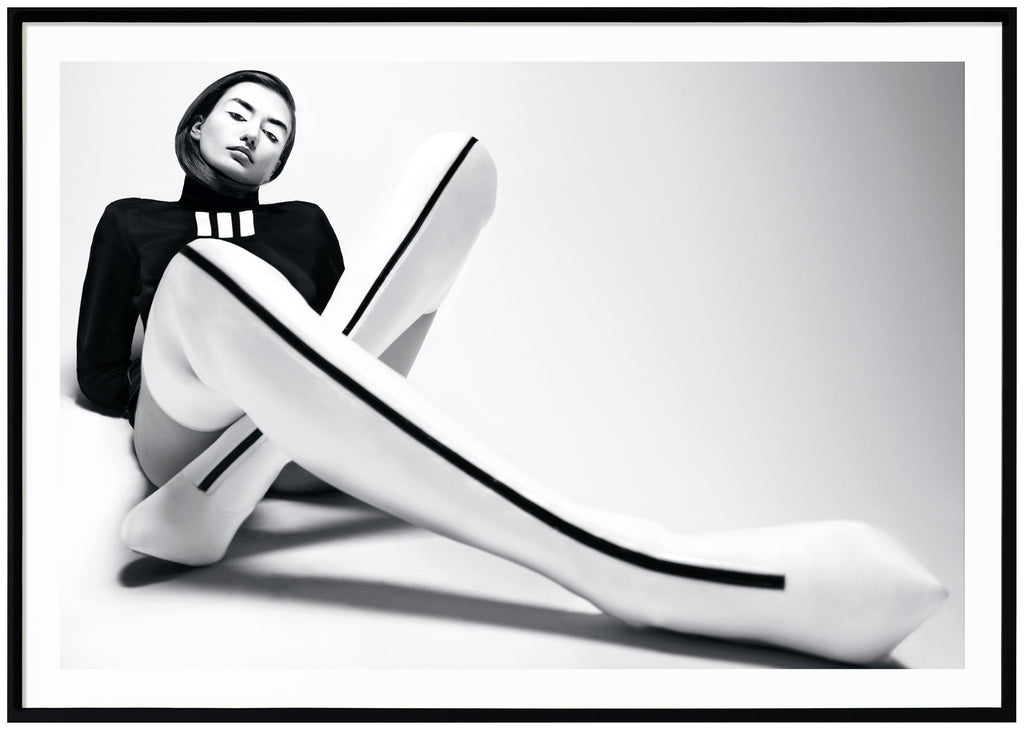 Svart-vit poster av kvinna sittandes på golvet. Vita overknee klackar med svart rand. Svart tröja. Liggande format. Svart ram.