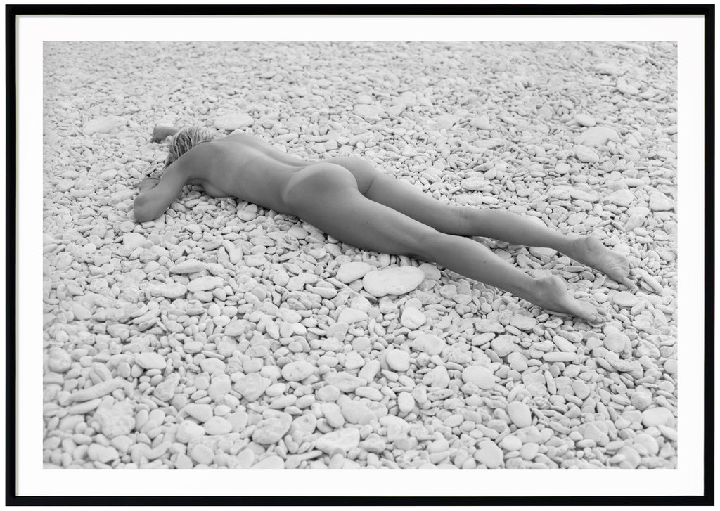 Svart-vit poster av kvinna liggandes på mage på strand med vita stenar. Liggande format. Svart ram.
