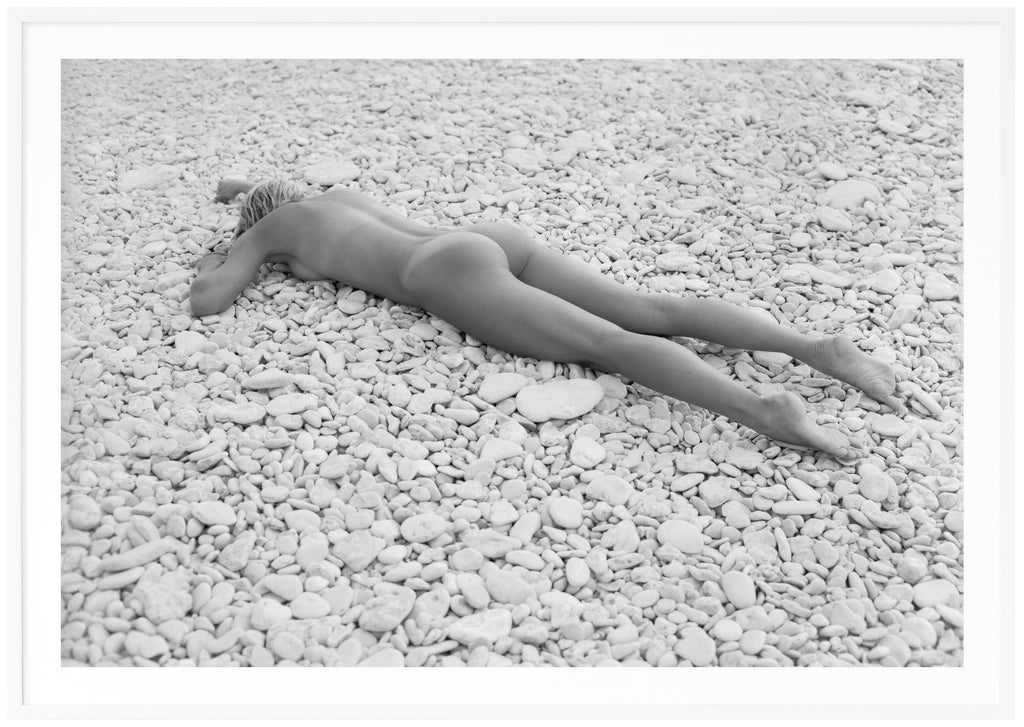 Svart-vit poster av kvinna liggandes på mage på strand med vita stenar. Liggande format. Vit ram.