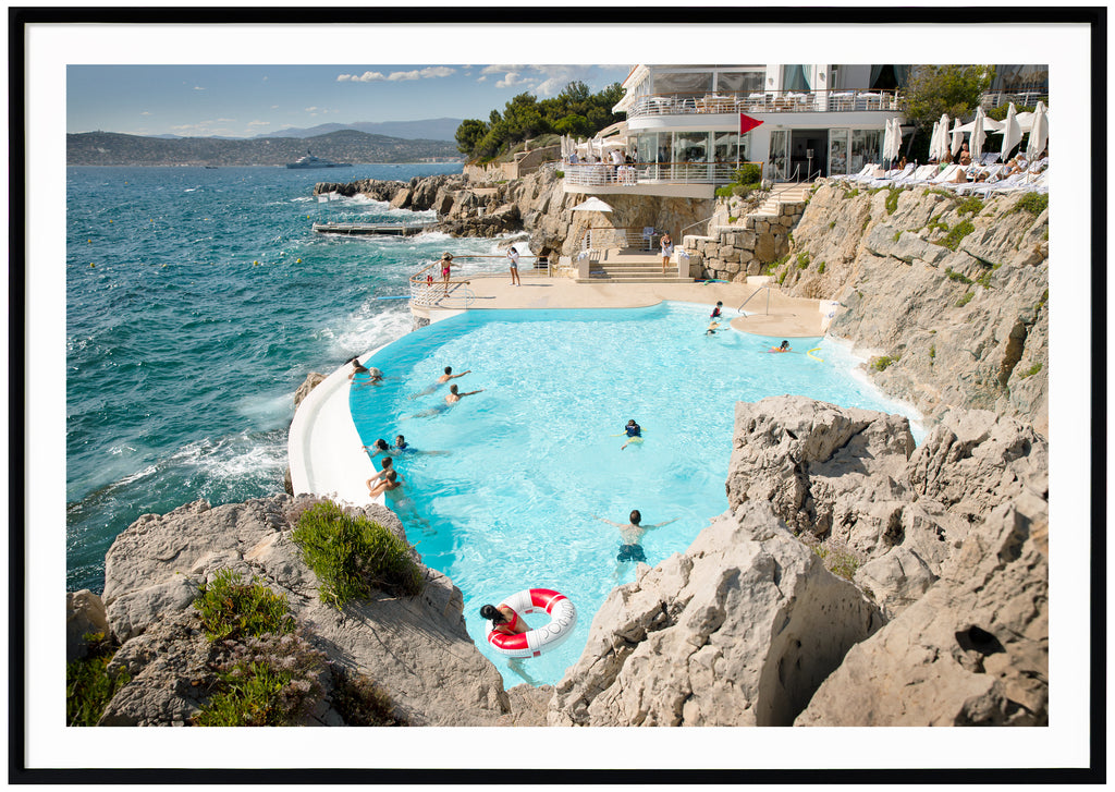 Poster av badande folk i pool vid franska rivieran. Vitt hotell med gröna träd bakom. Röd flagga uppe. Yacht i havet. Svart ram.