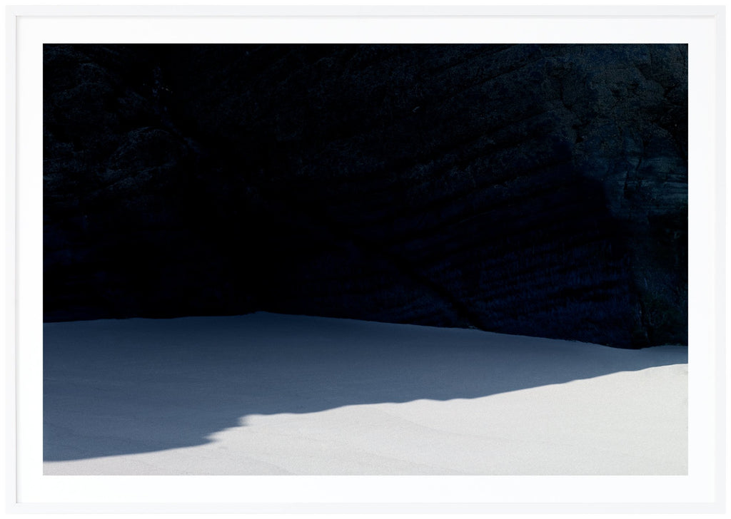 Grafiskt poster av en strand med en skugga från en mörk klippa. Liggande format. Vit ram.
