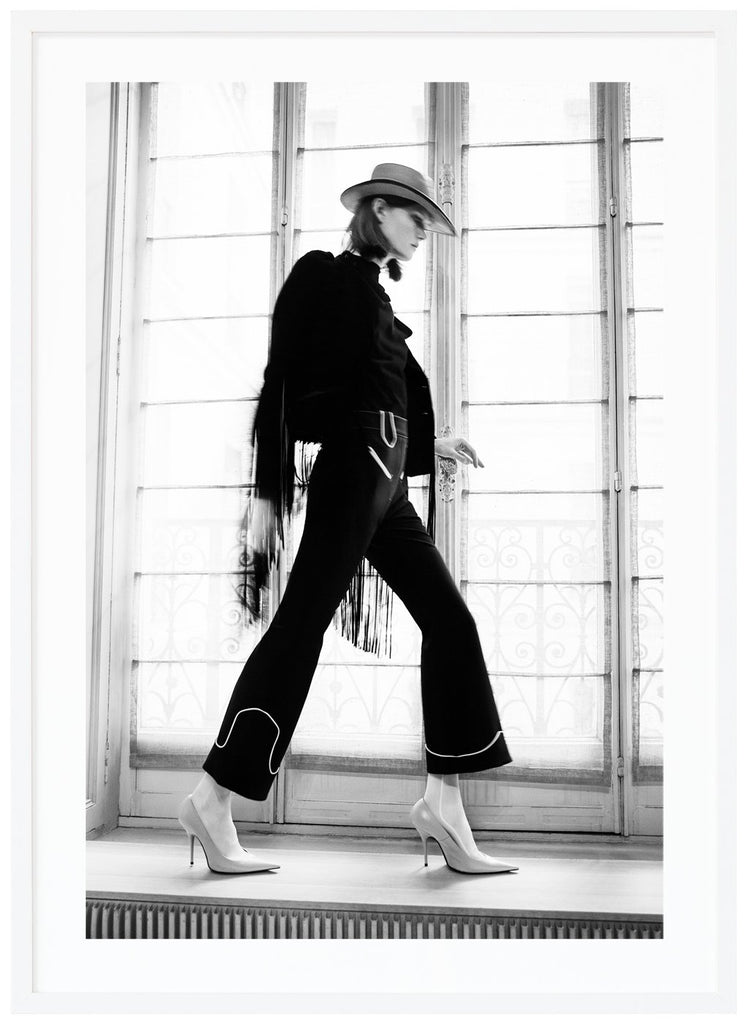 Svart-vit poster av kvinna framför stort fönster sedd från sidan med klackskor hatt och svart klädsel. Vit ram.