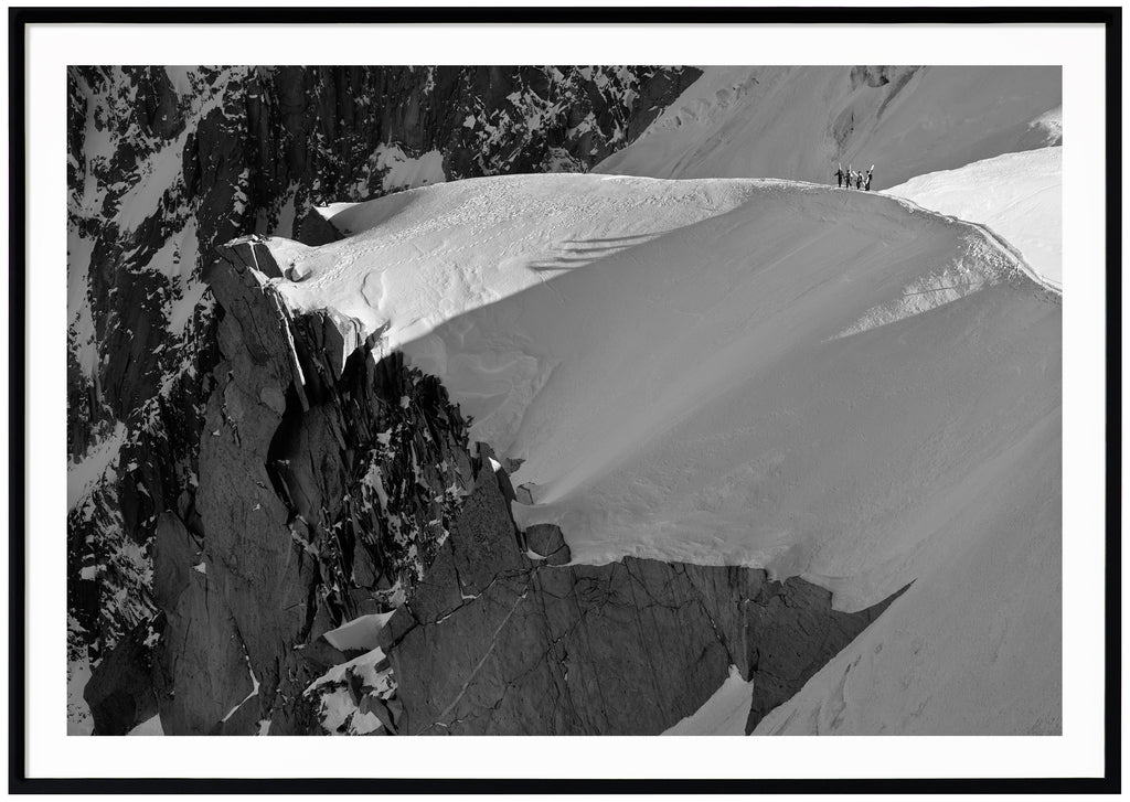 Svart-vitt fotografi av fyra skidåkare som vandrar på snötäckt bergskam. Svart ram