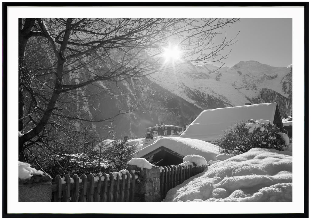Svart-vitt fotografi av snötäckta hustak med alper i bakgrunden. Svart ram
