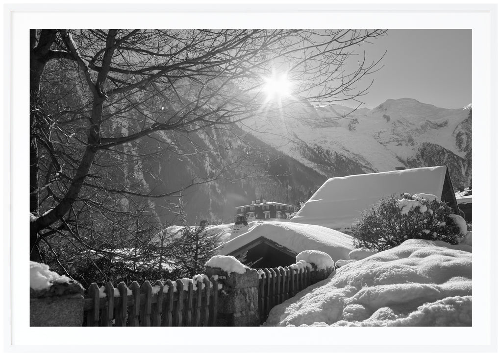 Svart-vitt fotografi av snötäckta hustak med alper i bakgrunden. Vit ram
