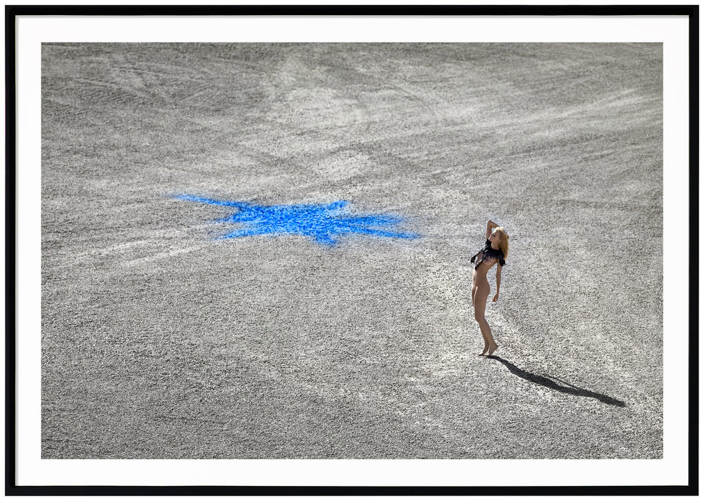Poster i liggande format av naken kvinna ståendes på stenmark med en blå figur på. Svart ram.