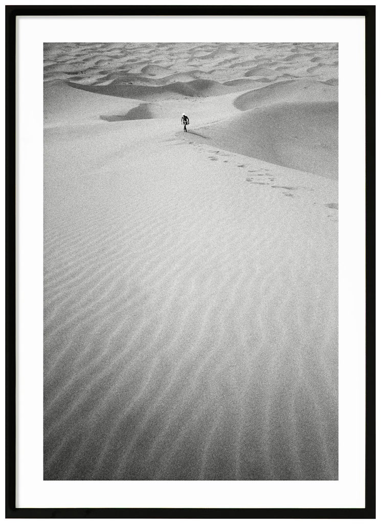 Svart-vitt analogt fotografi av en man på långt håll, som vandrar uppför en enorm sanddyna mitt i Saharaöknen. Svart ram.