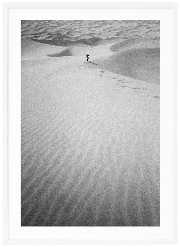 Svart-vitt analogt fotografi av en man på långt håll, som vandrar uppför en enorm sanddyna mitt i Saharaöknen. Vit ram.